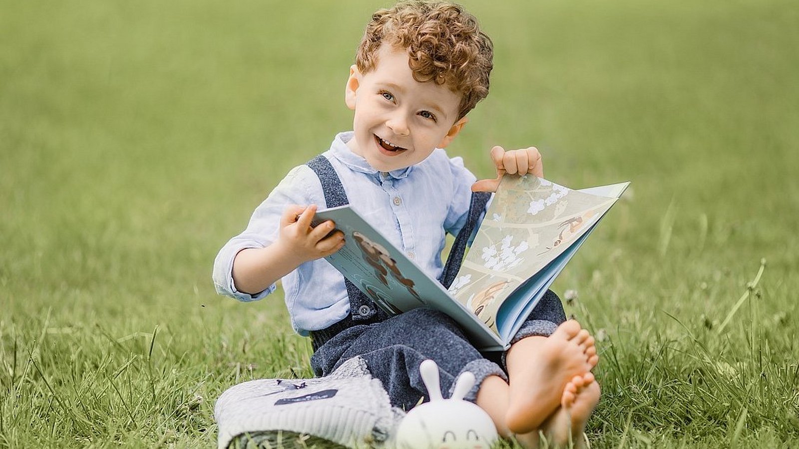 Ein Junge im Kita-Alter sitzt im Sommer auf einem grünen Rasen. Er hält ein geöffnetes Bilderbuch auf seinen gekreuzten Beinen und lacht. 