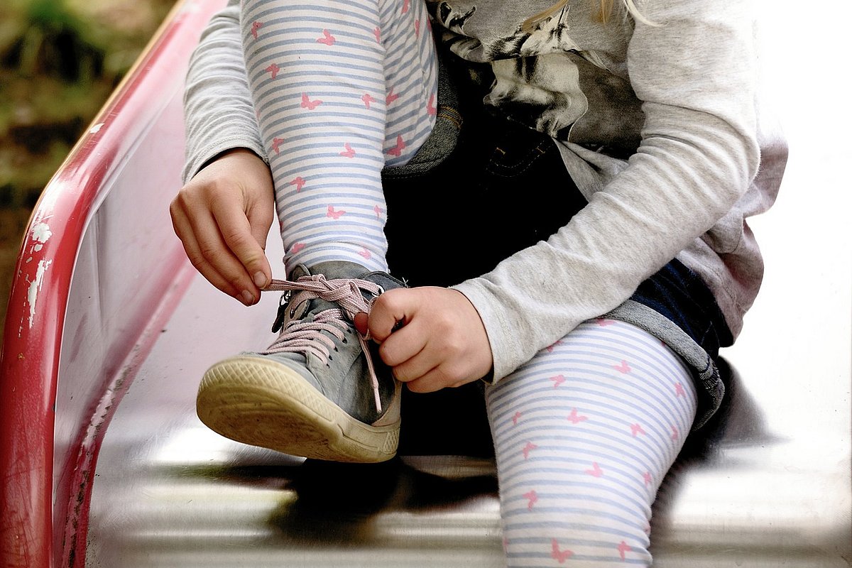 Ein Mädchen sitzt auf einer Spielplatzrutsche und bindet sich die Schnürsenkel zu. 