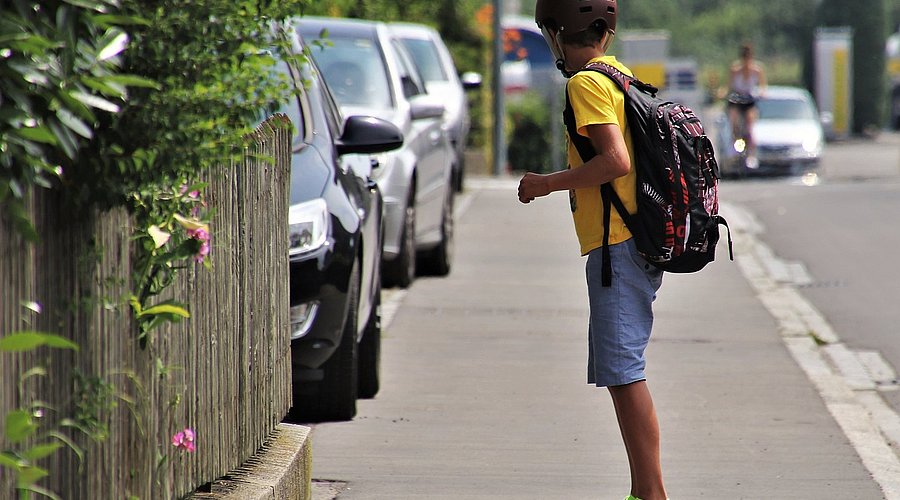 Ein Junge fährt auf einem Skateboard über den Bürgersteig. Er trägt kurze Hosen und Helm.