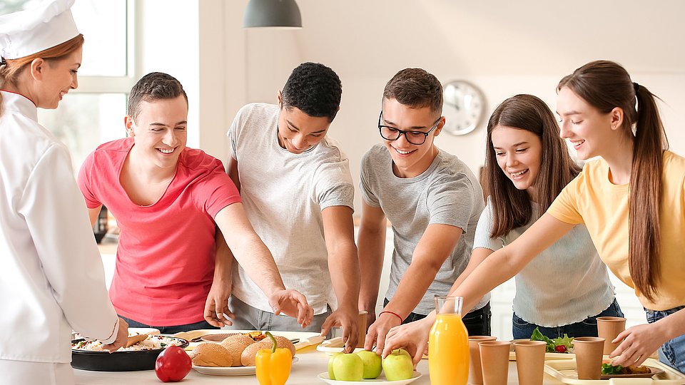 Auf einem weißen Tisch sind Speisen, Obst und Gemüse präsentiert. Davor stehen fünf junge Erwachsene mit ihren Tabletts, auf denen Essen steht. Sie greifen alle gemeinsam nach einem Teller mit grünen Äpfeln. 