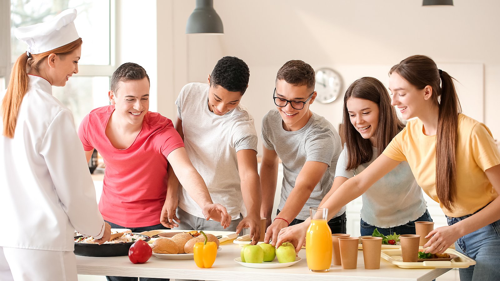 Auf einem weißen Tisch sind Speisen, Obst und Gemüse präsentiert. Davor stehen fünf junge Erwachsene mit ihren Tabletts, auf denen Essen steht. Sie greifen alle gemeinsam nach einem Teller mit grünen Äpfeln. 