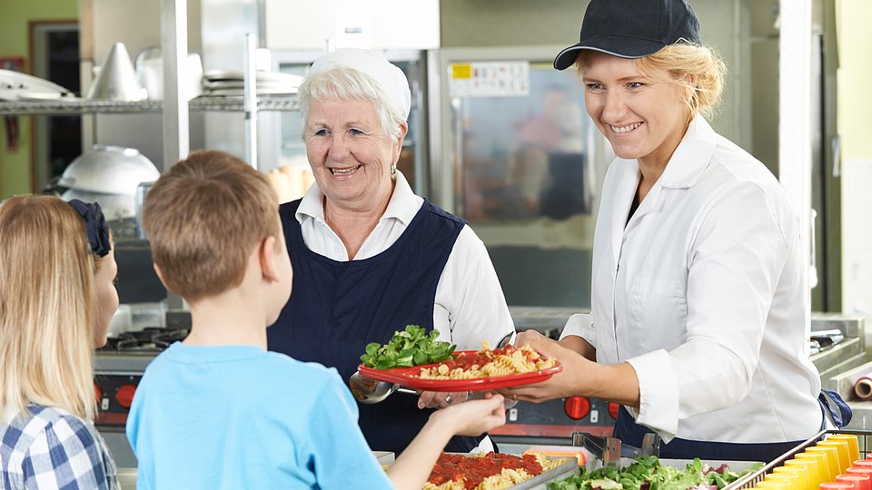 Zwei kinder stehen an der Ausgabetheke der Schulverfpelgung und erhalten Menü von freundlicher Servicekraft