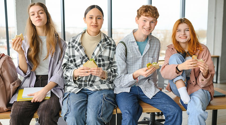 Teenager sitzen in der Schulmensa auf einem Tisch und snacken.