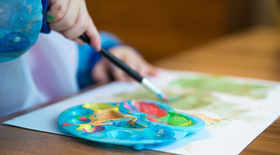Ein Kinder-Farbenpalette, eine Kinderhand mit Pinsel in der Hand und ein bemaltes Blatt Papier. Ein Kind sitzt am Tisch und malt.