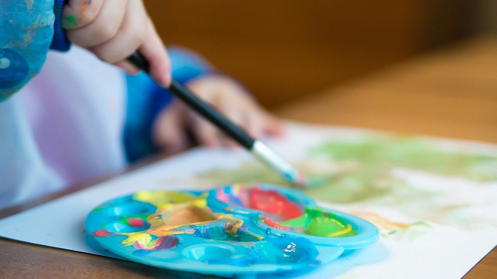 Ein Kinder-Farbenpalette, eine Kinderhand mit Pinsel in der Hand und ein bemaltes Blatt Papier. Ein Kind sitzt am Tisch und malt.