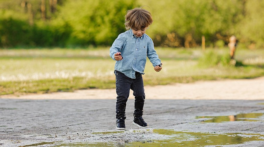 Ein Junge im Kindergartenalter springt in einer Pfütze auf und ab.