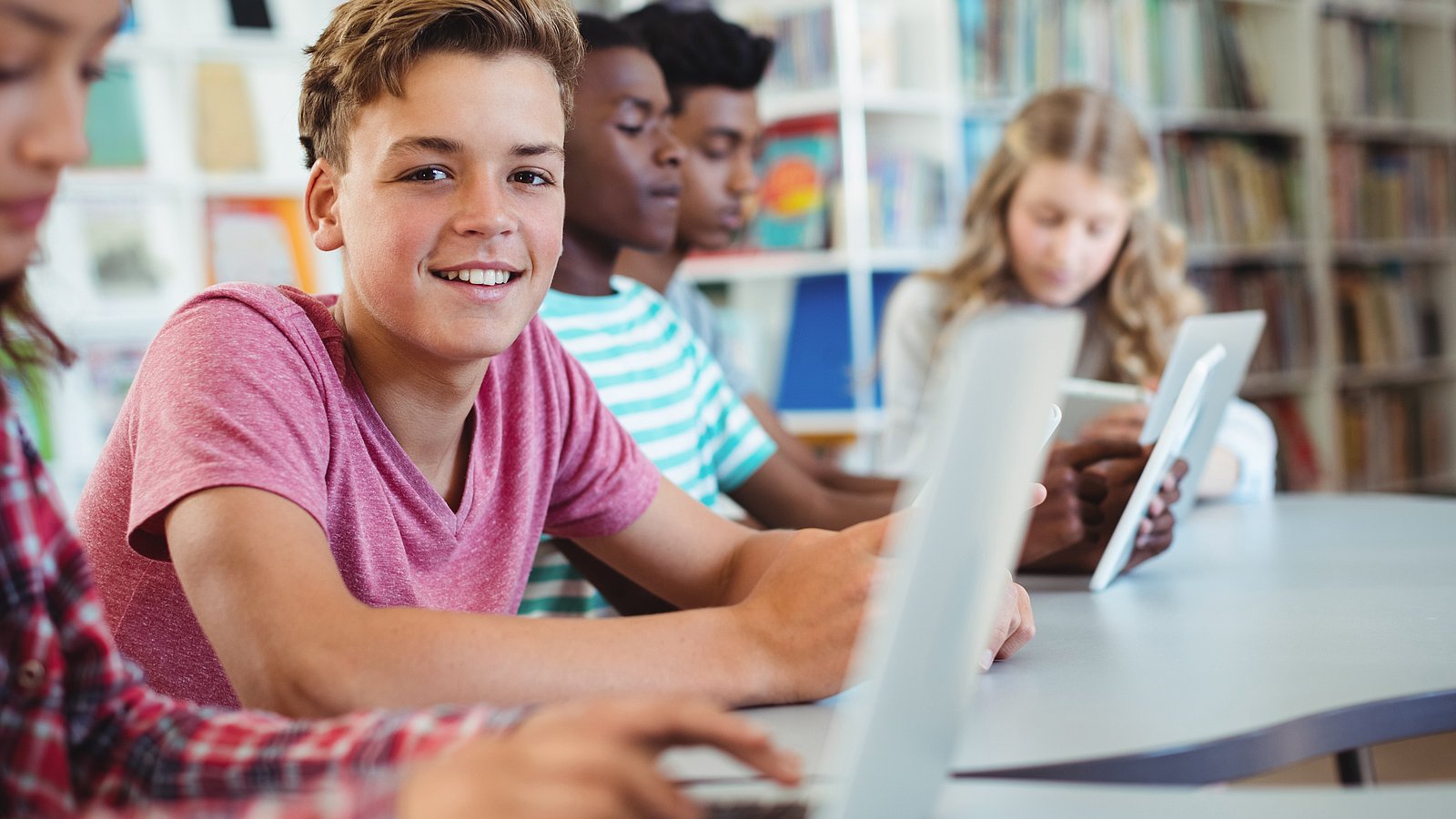 Jugendliche Schüler*innen sitzen in einer Schulbibliothek am Laptop. Ein Junge schaut lächelnd in die Kamera, alle anderen konzentrieren sich auf ihre Arbeit. 