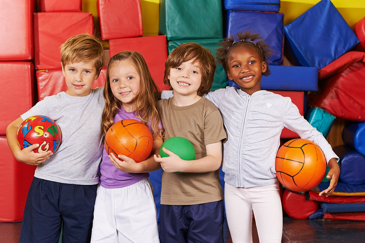 Vier Kinder mit unterschiedlicher Hautfarbe stehen untergehakt in einer Turnhalle vor bunten Sportmatten und halten jeweils einen Ball in der Hand. 