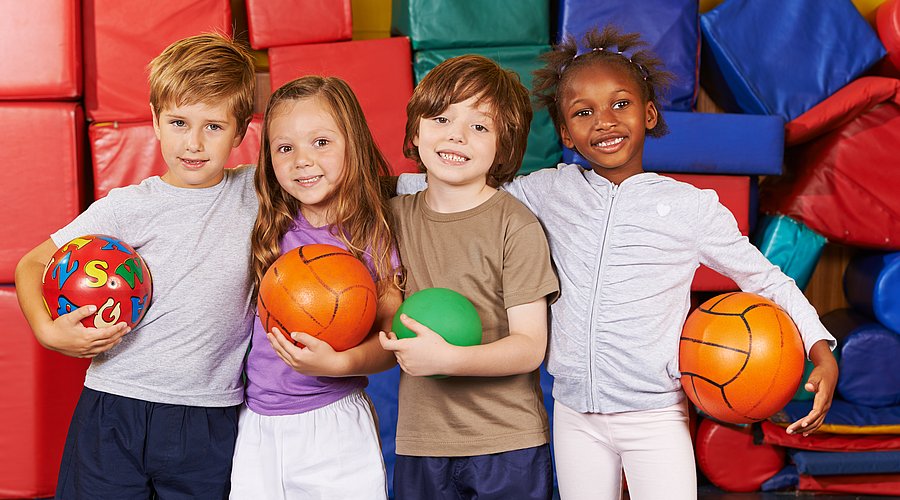 Vier Kinder mit unterschiedlicher Hautfarbe stehen untergehakt in einer Turnhalle vor bunten Sportmatten und halten jeweils einen Ball in der Hand. 