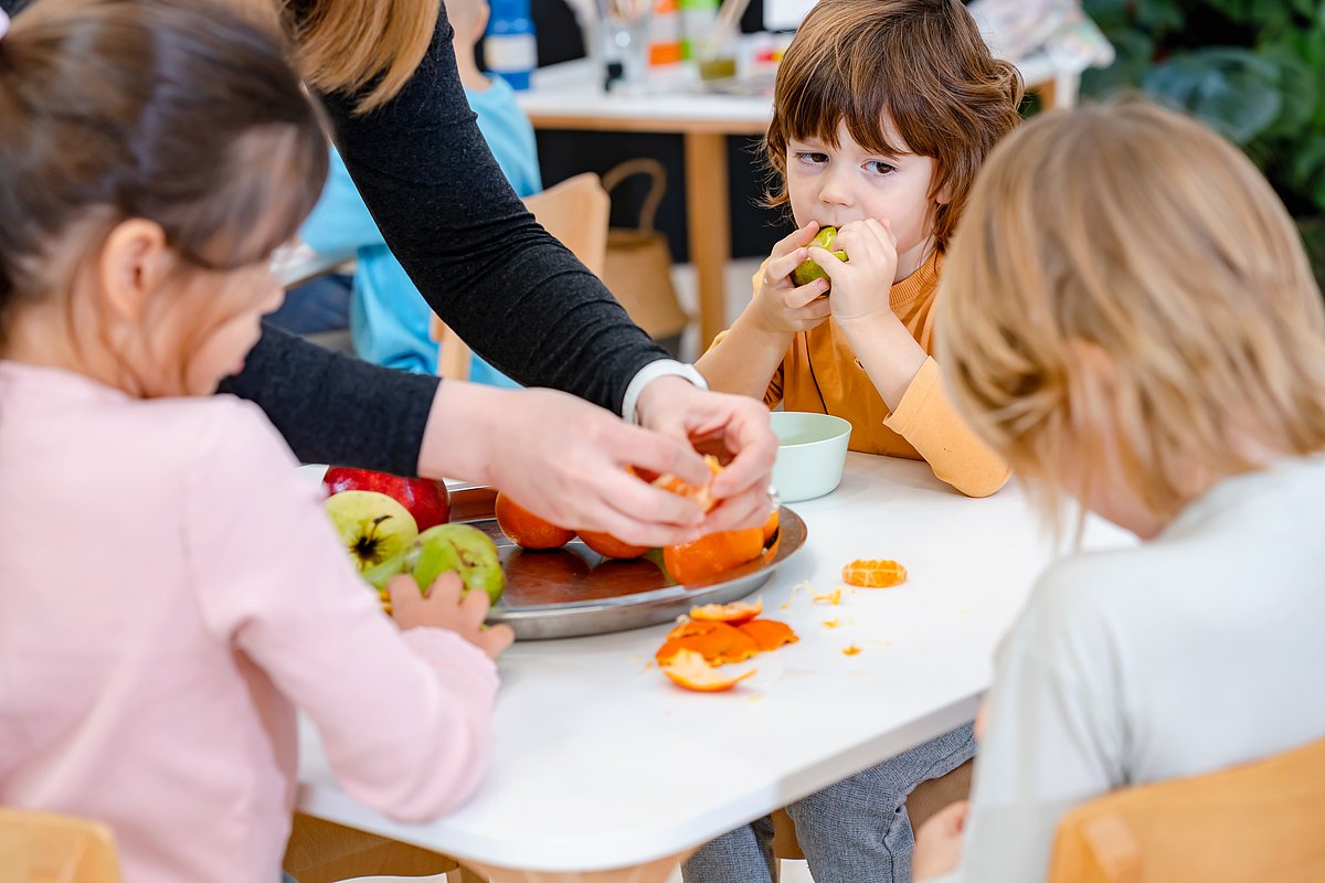 Kita-Kinder sitzen um einen Tisch und essen Obst von einer Obstplatte.